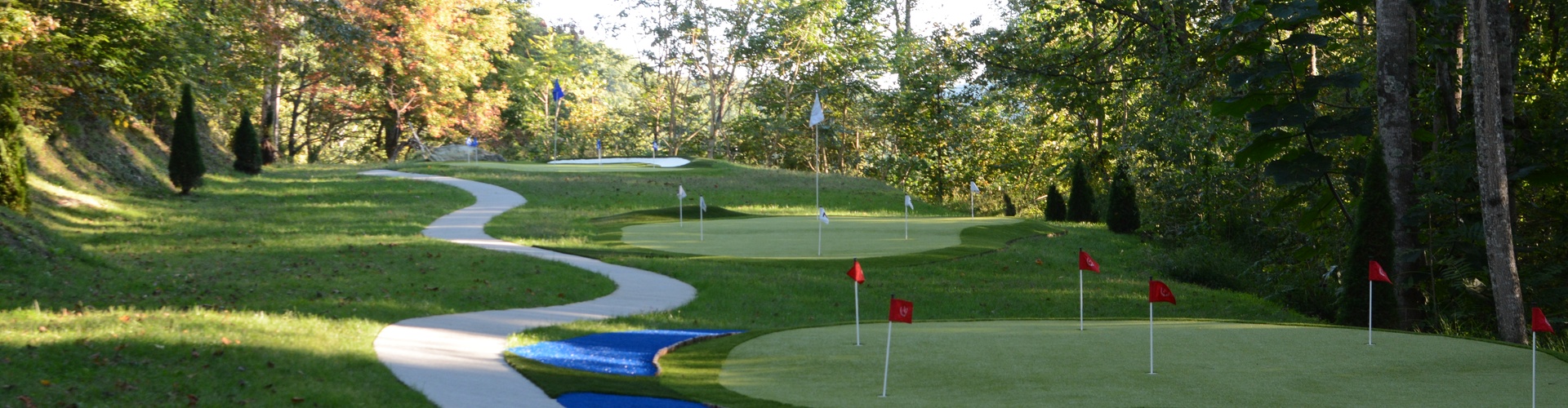 Artificial turf installation on a multi-hole residential golf course with red flag pins, a winding cart path, water feature, and mature hardwood tree canopy in fall foliage